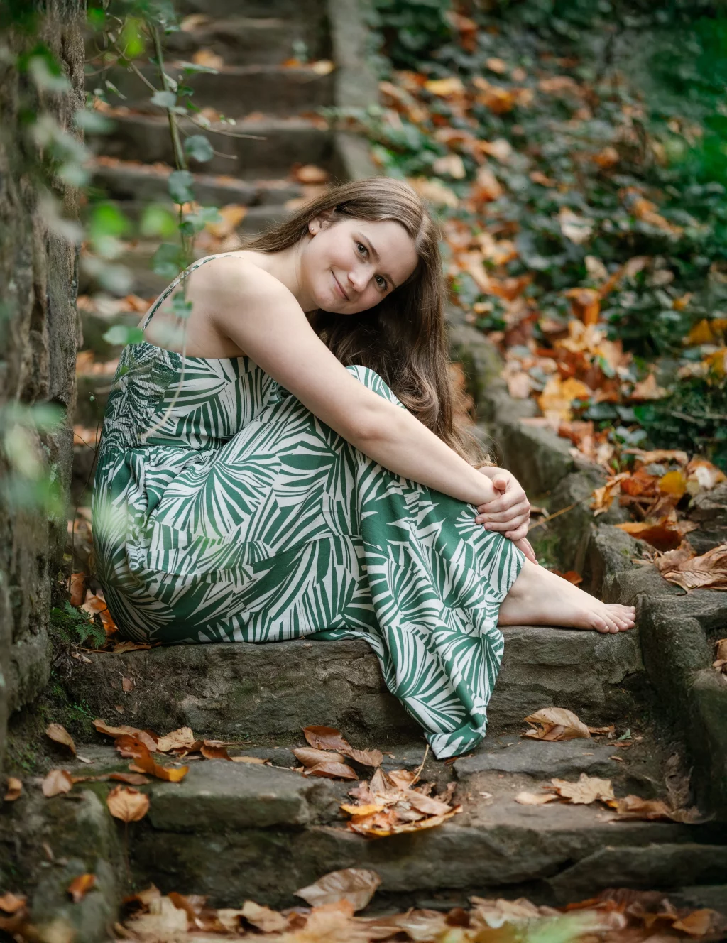 Sabrina's senior photos taken downtown Greenville, SC: Young woman in a green and white leaf-patterned dress sitting barefoot on outdoor stone steps surrounded by autumn leaves, smiling at the camera.