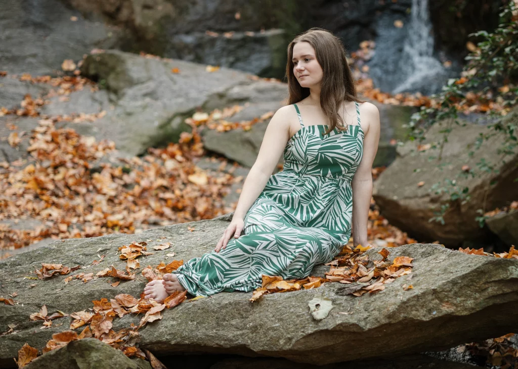 Sabrina's senior photos taken downtown Greenville, SC: Woman in a green and white patterned dress sitting barefoot on a rock surrounded by fallen autumn leaves, with a waterfall in the background.