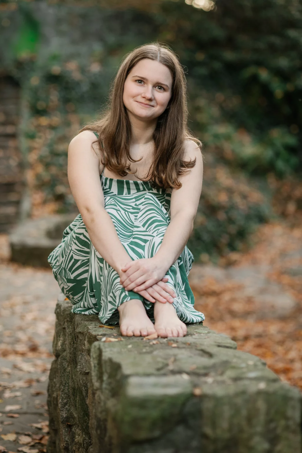 Sabrina's senior photos taken downtown Greenville, SC: Young woman with long brown hair sitting barefoot on a stone wall outdoors, wearing a green and white patterned dress, surrounded by fallen leaves.