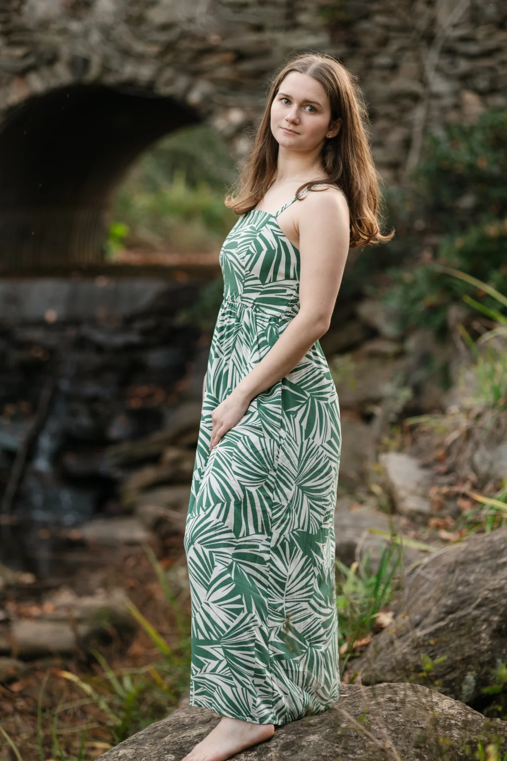 Sabrina's senior photos taken downtown Greenville, SC: Young woman in a green and white patterned maxi dress standing barefoot on a rock near a stone bridge and greenery.