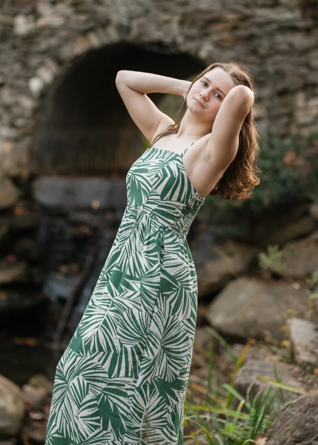 Sabrina's senior photos taken downtown Greenville, SC: Young woman in a green and white leaf-patterned dress posing outdoors with hands behind her head, standing in front of a stone bridge and rocks.