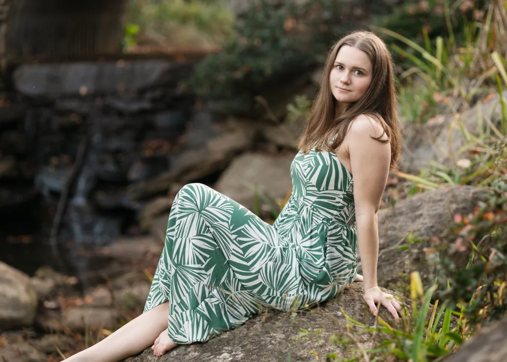 Sabrina's senior photos taken downtown Greenville, SC: Young woman in a green and white leaf-patterned dress sitting barefoot on a large rock near a natural outdoor setting with blurred foliage and rocks in the background.