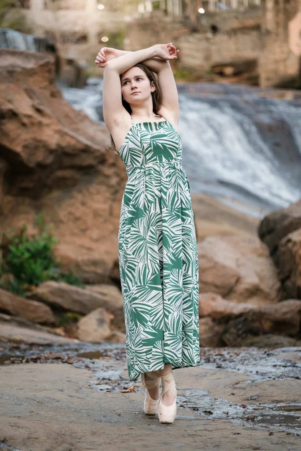 Sabrina's senior photos taken downtown Greenville, SC: Young woman in a green and white leaf-patterned dress and ballet pointe shoes standing on rocky ground near a flowing waterfall, arms raised above her head.