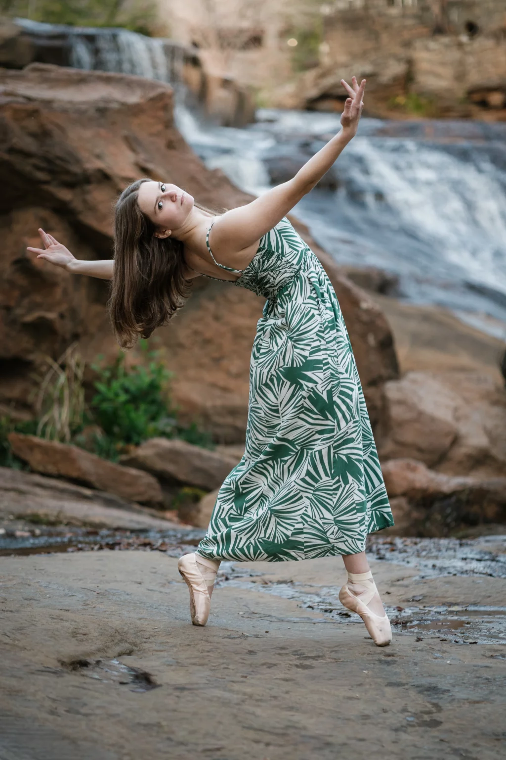 Sabrina's senior photos taken downtown Greenville, SC: Female ballet dancer in green and white dress posing en pointe on rocky ground with waterfall in the background.