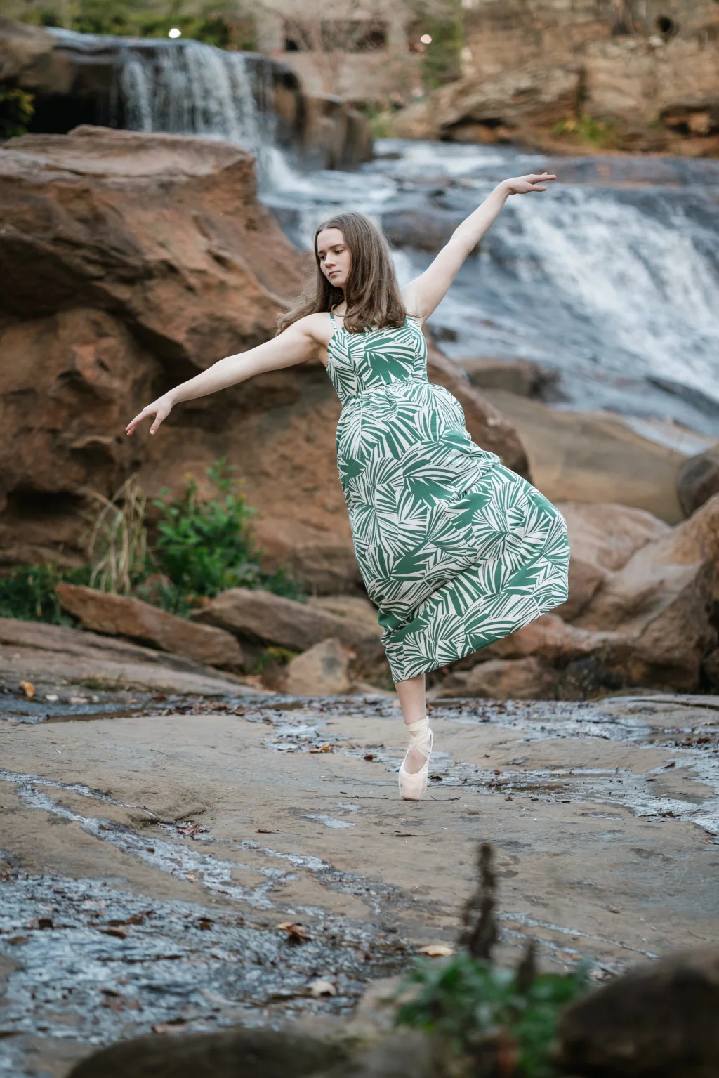 Sabrina's senior photos taken downtown Greenville, SC: Young woman in a green and white patterned dress dancing on pointe shoes on rocks near a waterfall.