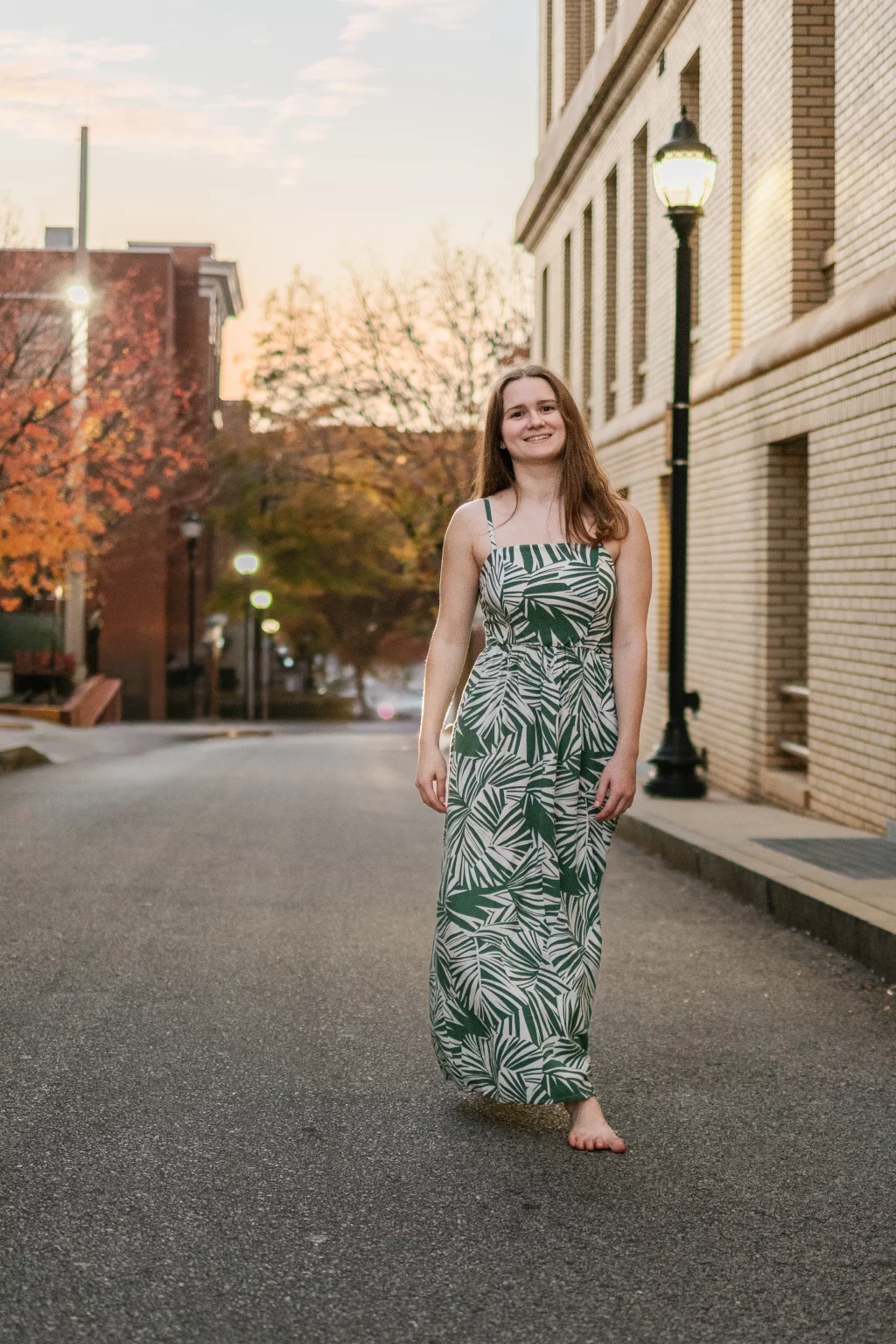 Sabrina's senior photos taken downtown Greenville, SC: Young woman smiling barefoot walking on empty street at dusk wearing a green and white leaf-patterned maxi dress.