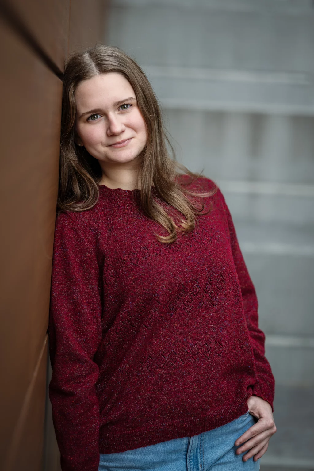Sabrina's senior photos taken downtown Greenville, SC: Young woman with long brown hair wearing a red knit sweater and blue jeans, leaning against a brown wall, smiling softly.