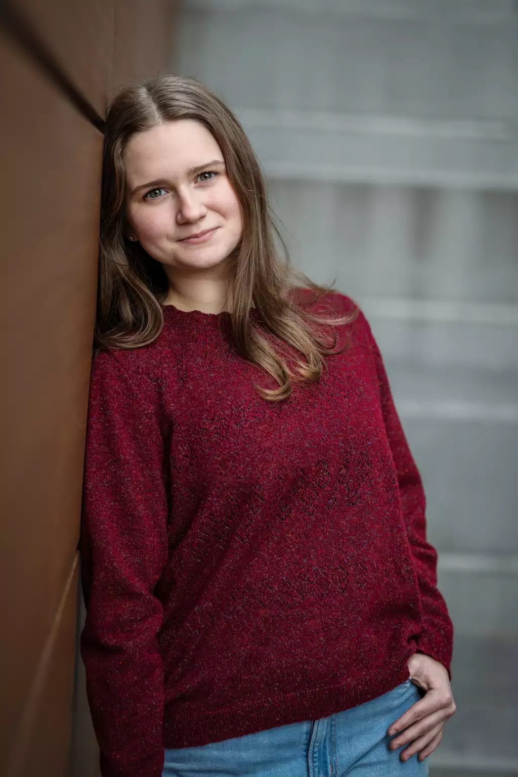 : Young woman with long brown hair wearing a red knit sweater and blue jeans, leaning against a brown wall, smiling softly.