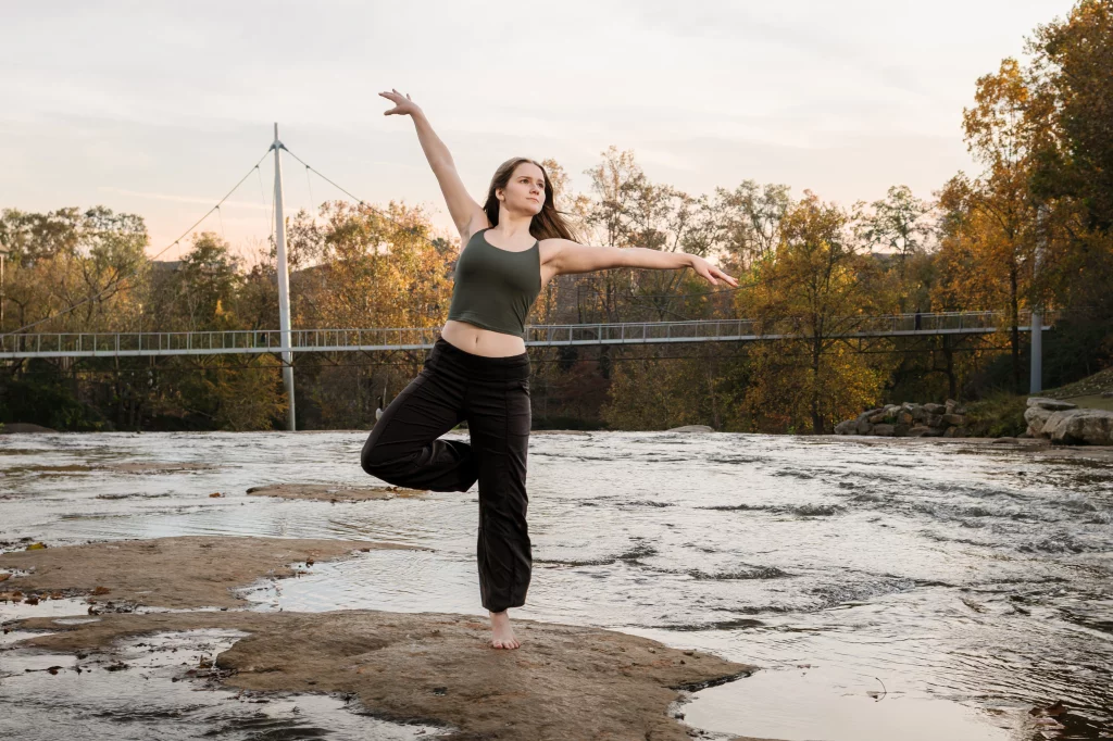 Sabrina's senior photos taken downtown Greenville, SC: Young woman in a green tank top and black pants balancing on one leg on a rock by a river with a pedestrian bridge and autumn trees in the background.