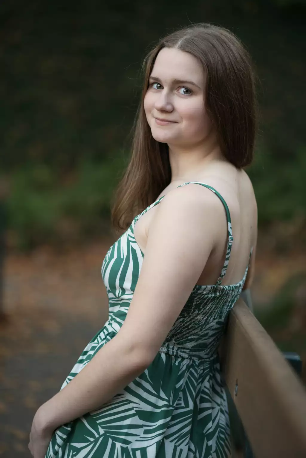 : Young woman with long brown hair sitting on a wooden bench outdoors, wearing a green and white patterned sleeveless dress, looking over her shoulder and smiling softly.