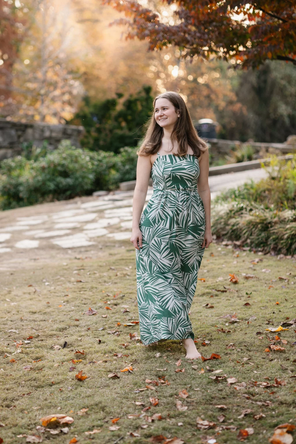 Sabrina's senior photos taken downtown Greenville, SC: Young woman in a green and white leaf-patterned maxi dress walking barefoot on grass with autumn leaves, smiling, with trees and a stone pathway in the background.