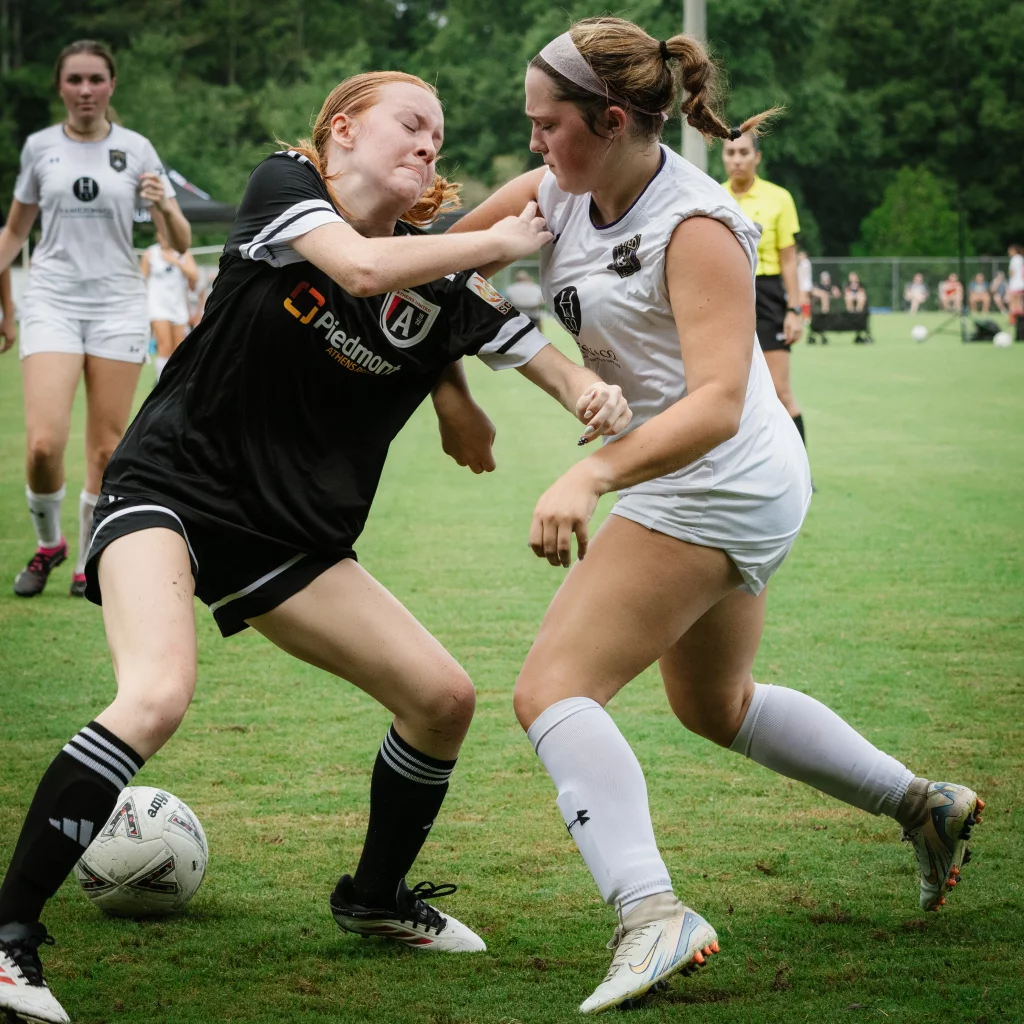 United FC - Furman Region -: Two female soccer players struggle for possession of the ball, with one player in a black uniform pushing against another player in a white uniform. Other players and a referee are visible in the background on a grassy field.