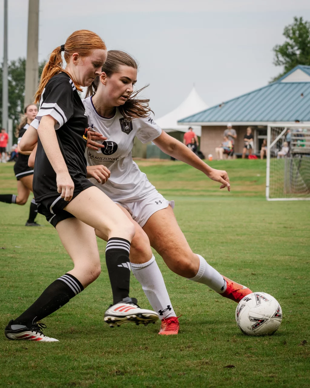 United FC - Furman Region -: Two teenage girls compete for the soccer ball during a match on a grassy field, one in a black uniform and the other in white, with a goal and building visible in the background.