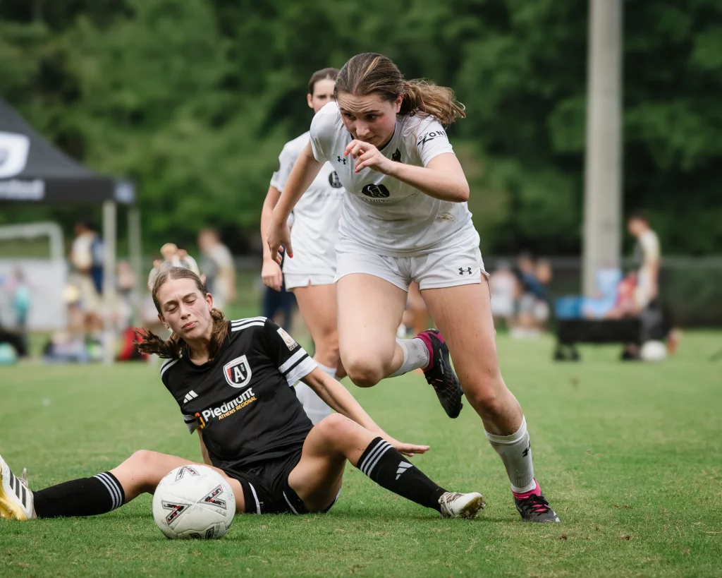United FC - Furman Region -: A soccer player in a white uniform runs with determination while a player in a black uniform slides on the grass attempting to take the ball.