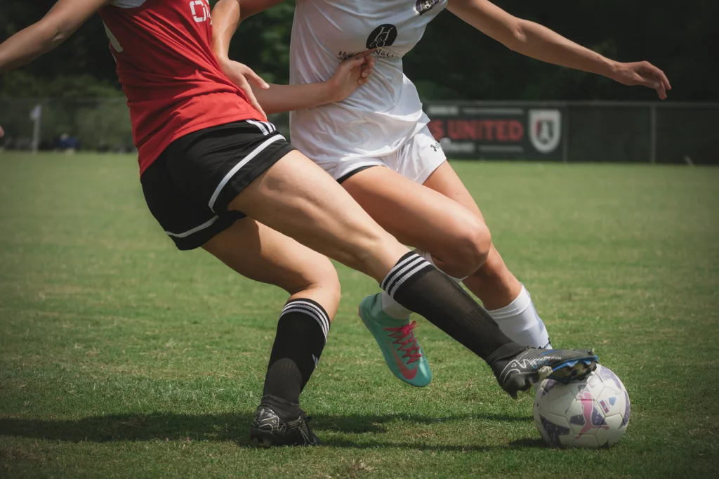 United FC - Furman Region -: Two soccer players competing for the ball on a grassy field, with only their legs and lower bodies visible. One wears a red jersey and black shorts, the other a white uniform.
