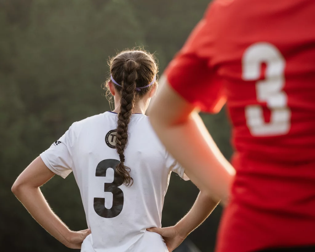 United FC - Furman Region -: Two soccer players with the number 3 on their jerseys, one in white with a braid and the other in red, stand on a field with hands on their hips.