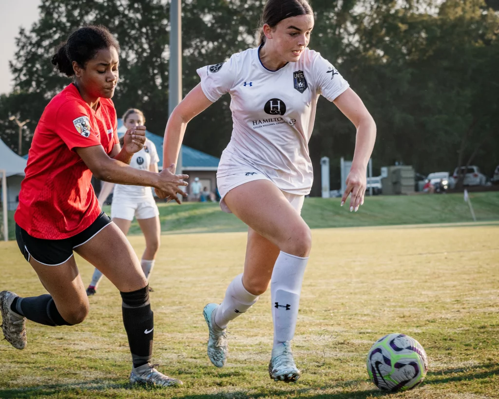 United FC - Furman Region -: Two female soccer players compete for the ball on a grassy field, one in a red jersey and the other in a white jersey, with other players and trees in the background.