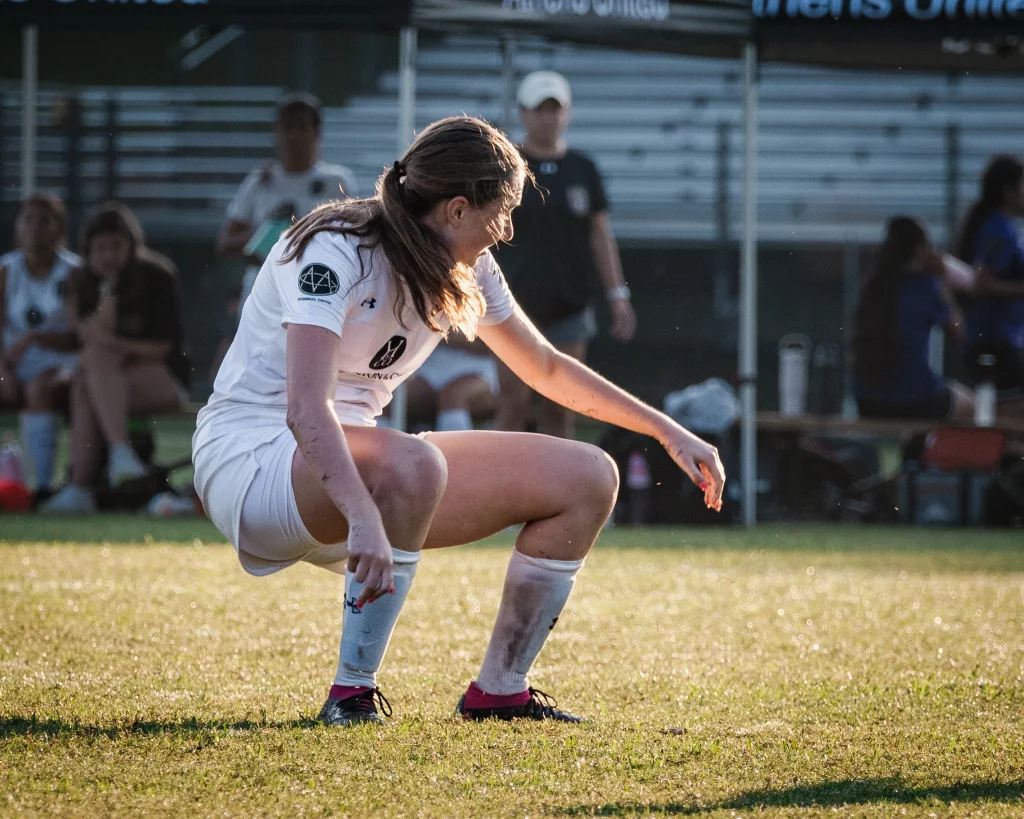 United FC - Furman Region -: A soccer player in a white uniform crouches on a grassy field, with sunlight highlighting her hair and arm. Teammates and spectators sit on benches in the blurred background.