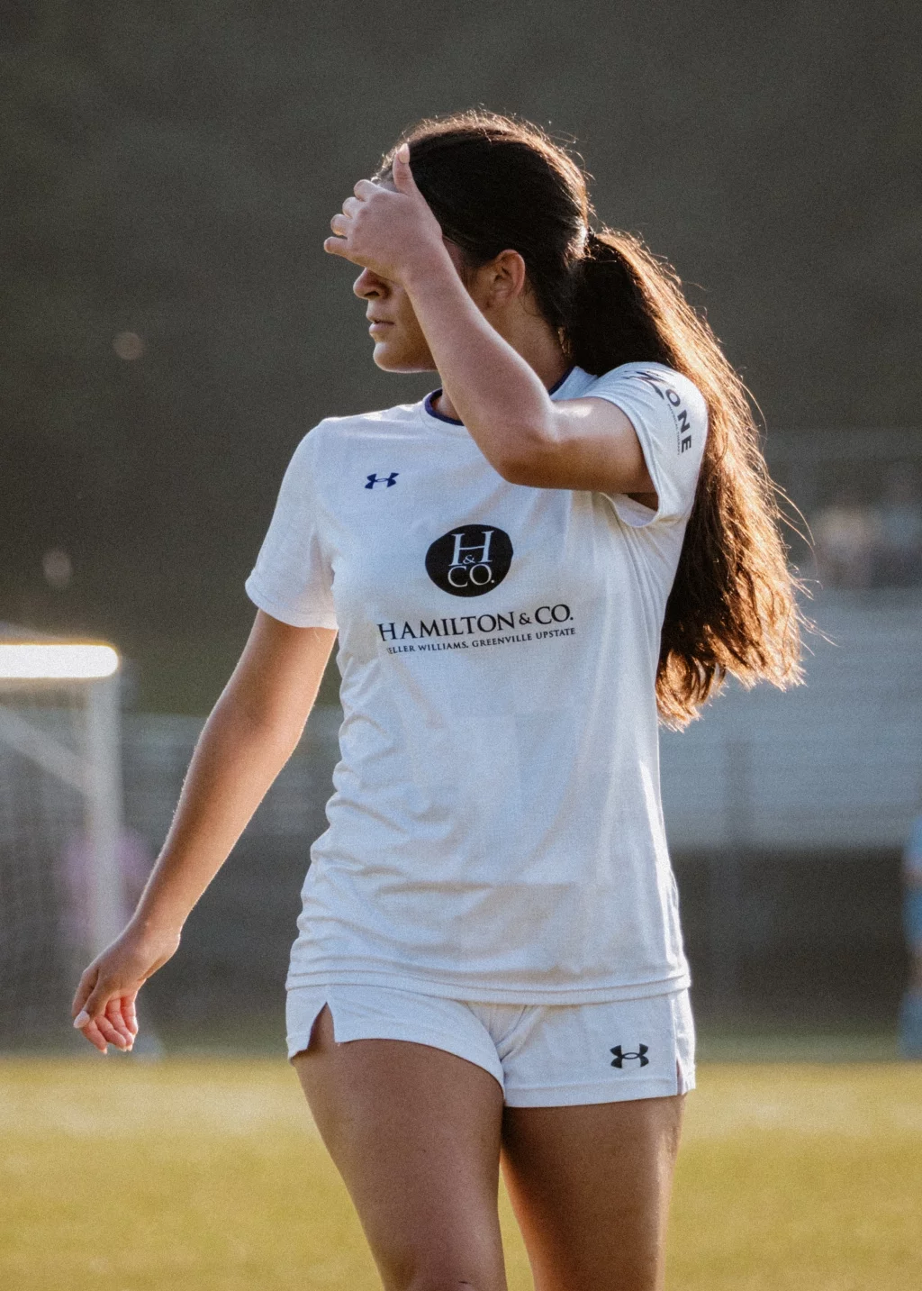 United FC - Furman Region -: A female soccer player in a white uniform stands on a field, shielding her eyes from the sun with her hand.