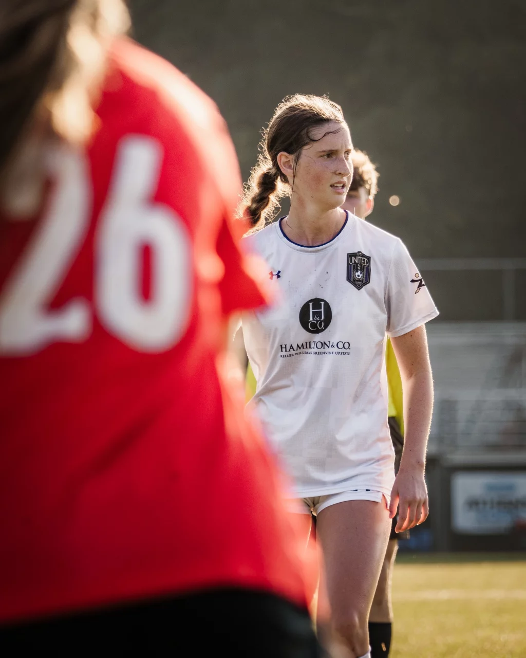 United FC - Furman Region -: A female soccer player in a white uniform stands on a sunlit field, looking to the side, with another player in a red jersey partially visible in the foreground.