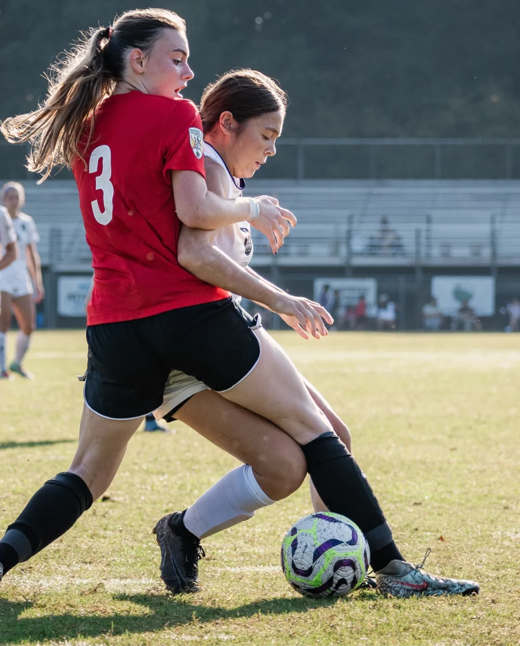 United FC - Furman Region -: Two female soccer players compete for the ball, with one in a red jersey and the other in a white jersey, closely contesting possession on a grassy field. Other players and spectators are visible in the background.