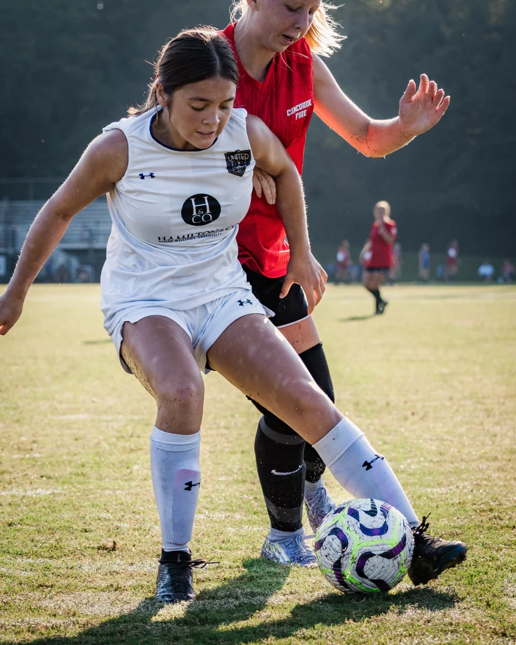 United FC - Furman Region -: Two female soccer players compete for the ball on a grassy field; one wears a white uniform and the other a red jersey.