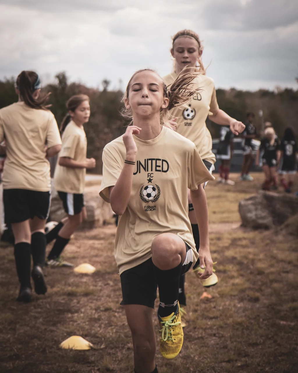 United FC - Furman Region -: Girls in yellow soccer uniforms running drills outdoors, with one girl in the foreground lifting her knee high.
