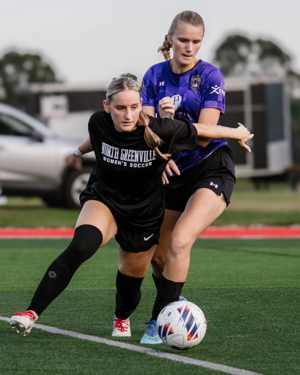 United FC - Furman Region -: Two female soccer players compete for the ball on a field; one in a black "North Greenville Women's Soccer" jersey, the other in a purple jersey, both in action near the sideline.