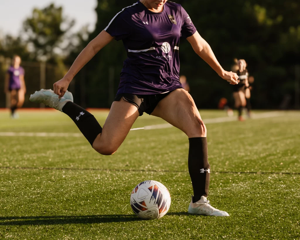 United FC - Furman Region -: Close-up of a soccer player in a purple jersey and black shorts preparing to kick a soccer ball on a field, with other players blurred in the background.