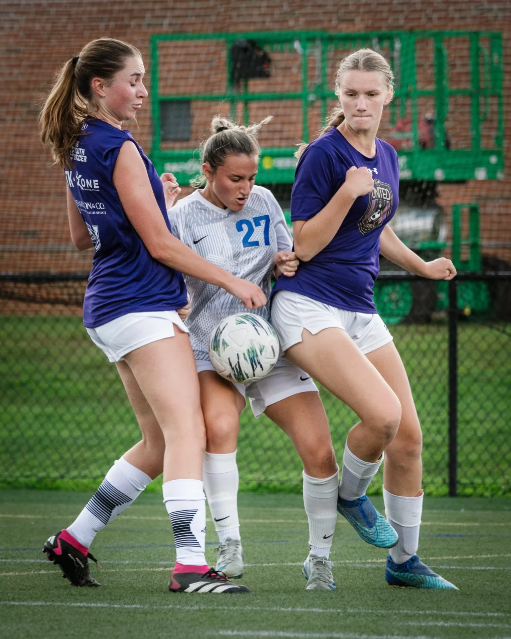 United FC - Furman Region -: Three female soccer players compete closely for the ball, with two players in blue shirts and one in a white shirt marked with the number 27, all focused on the ball at their feet on a turf field.