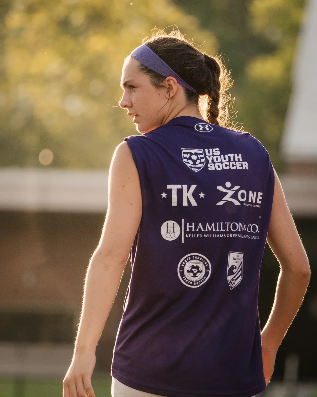 United FC - Furman Region -: A young soccer player with a purple headband and braided hair stands outdoors, wearing a sleeveless purple shirt with various logos including "US Youth Soccer" on the back. Sunlight highlights her hair, and the background is softly blurred.
