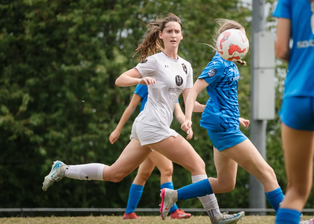 United FC - Furman Region -: A soccer player in white runs alongside players in blue, while a soccer ball obscures the face of the player in blue closest to the camera.