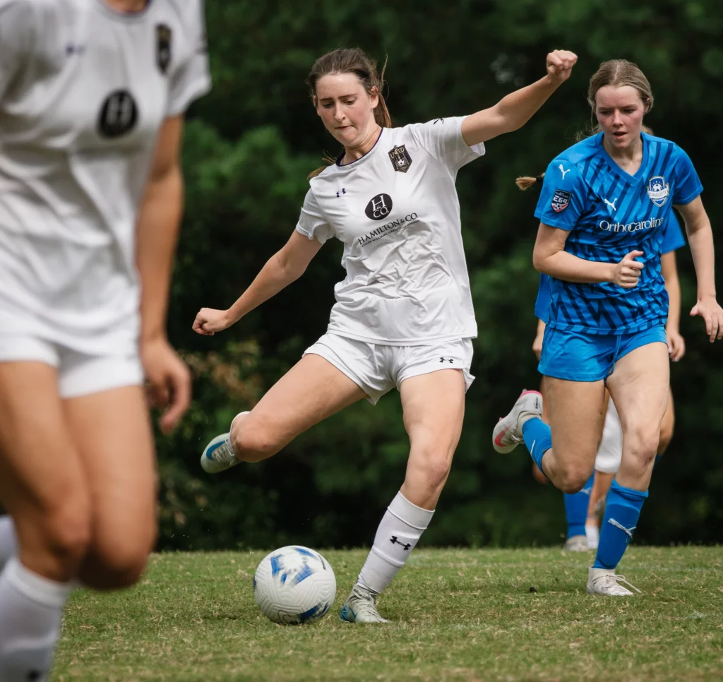 United FC - Furman Region -: A female soccer player in a white uniform kicks a ball while another player in a blue uniform runs nearby on a grassy field.
