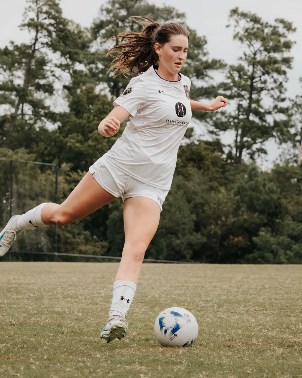 United FC - Furman Region -: Soccer player in a white uniform kicking a ball on a grassy field with trees in the background.
