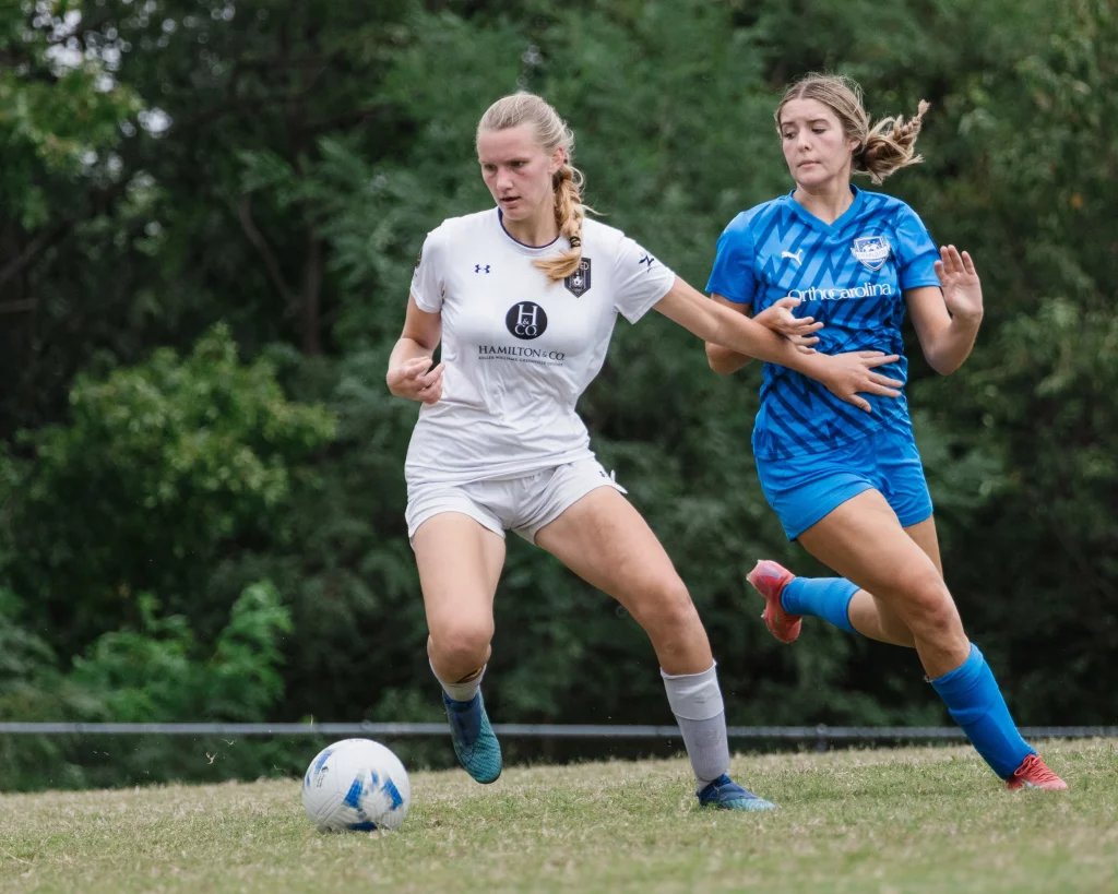 United FC - Furman Region -: Two female soccer players compete for the ball on a grassy field; one in a white uniform is advancing with the ball while the other in blue attempts to defend. Trees are visible in the background.