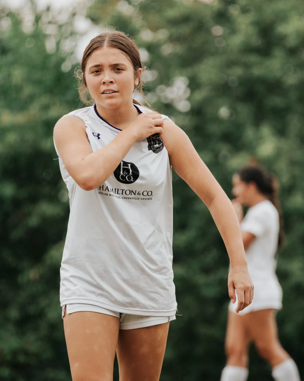 United FC - Furman Region -: A young female soccer player in a white uniform stands outdoors, touching her shoulder, with another player in the background. Trees are visible behind them.