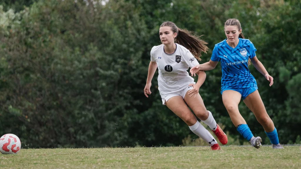 United FC - Furman Region -: Two female soccer players in action on a field, one in a white uniform and the other in a blue uniform, both running toward a soccer ball with trees in the background.