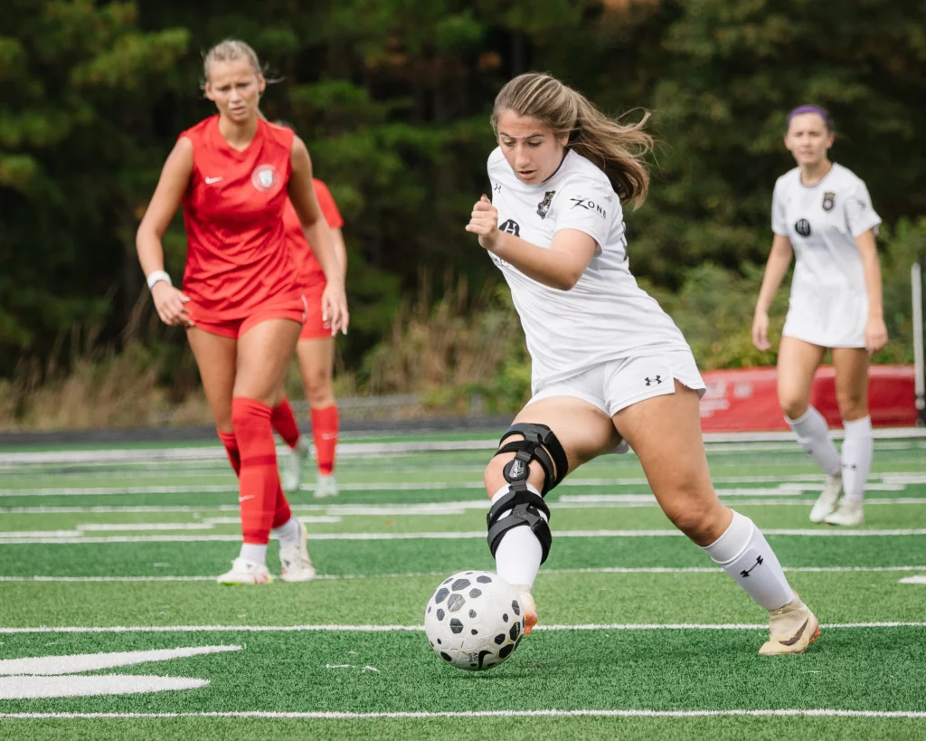 United FC - Furman Region -: A female soccer player in a white uniform with a knee brace is about to kick a soccer ball during a game, with opponents in red uniforms and teammates in white visible in the background on a green field.