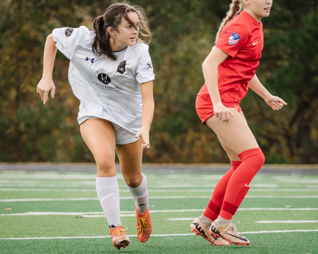 United FC - Furman Region -: Two young female soccer players in action on a field, one in a white uniform and the other in a red uniform, both focused and moving quickly with blurred trees in the background.