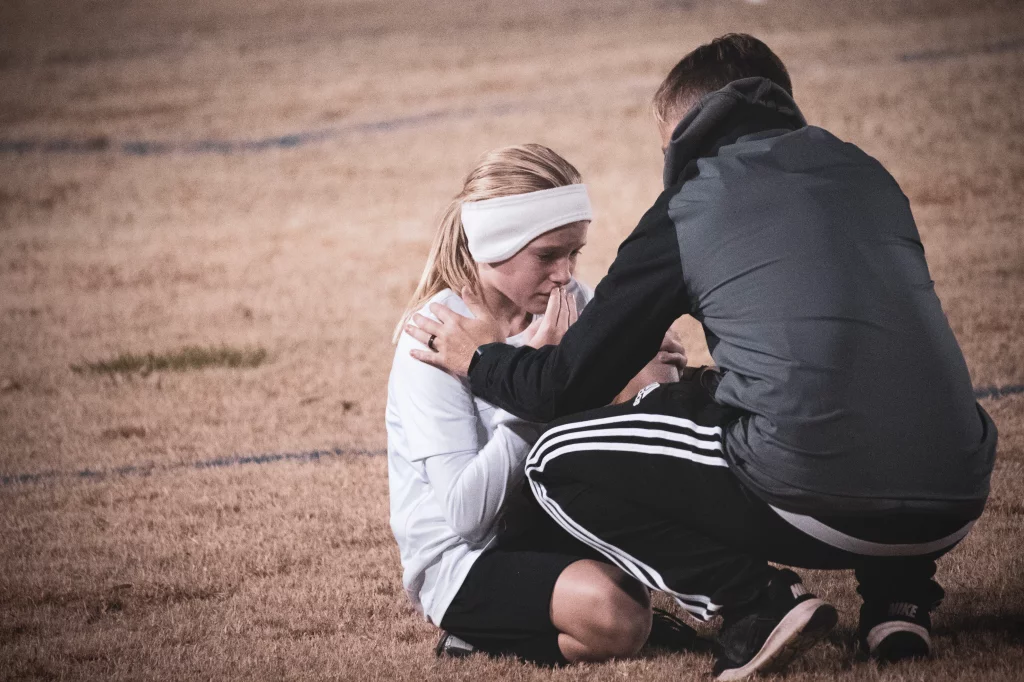 United FC - Furman Region -: A young soccer player sits on the field looking upset while an adult, possibly a coach, kneels beside her offering comfort with a hand on her shoulder.