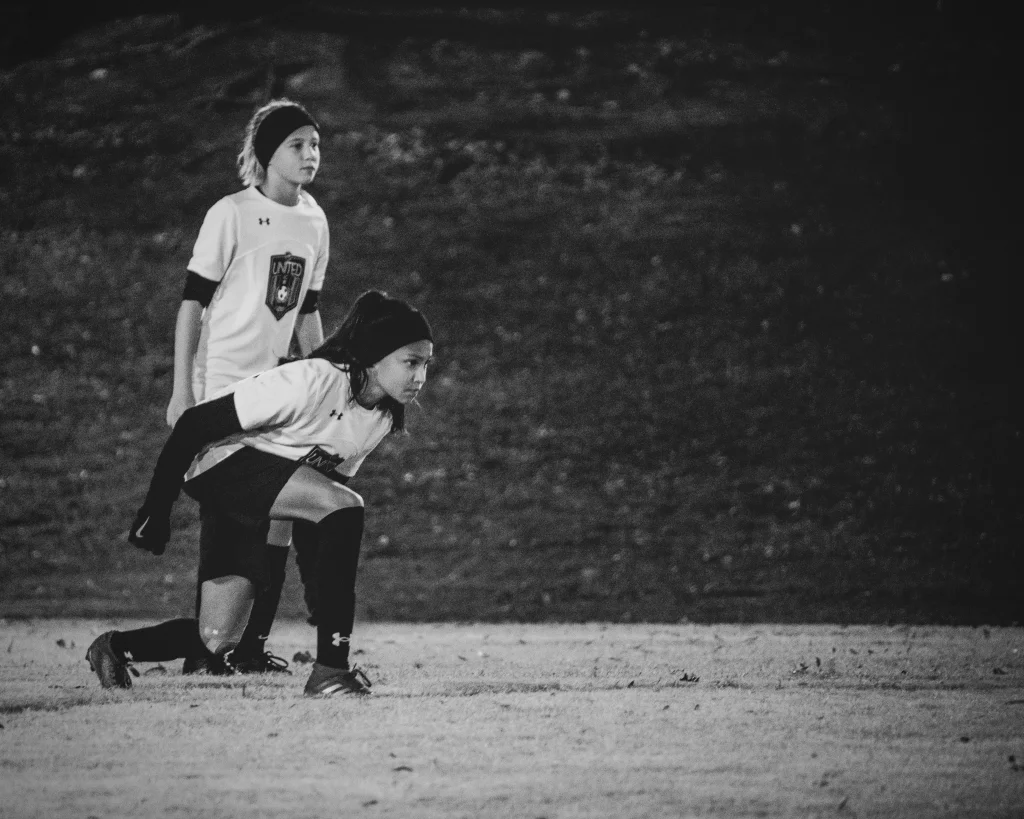 : Two young soccer players in uniform, one crouched and ready to move, the other standing behind, both focused on the game. The image is in black and white.