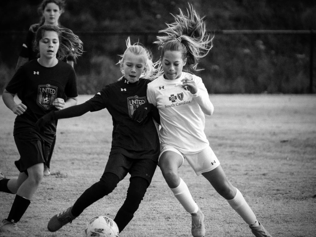 : Black and white photo of girls playing soccer, with two players in the foreground competing for the ball&mdash;one in a dark "United" uniform and the other in a white "South Carolina" uniform. Two more players in dark uniforms are visible in the background.