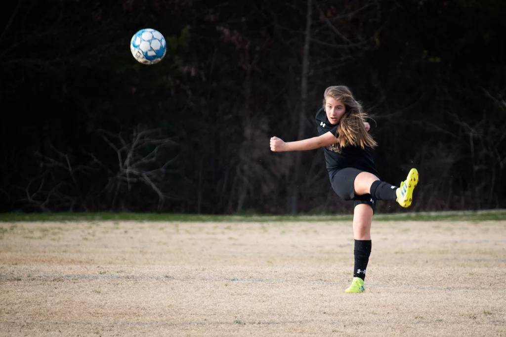 United FC - Furman Region -: A girl in a black soccer uniform kicks a blue and white soccer ball on a grassy field with trees in the background.
