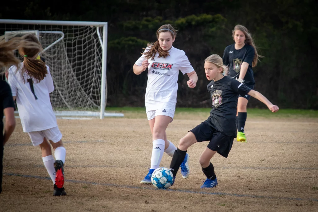 United FC - Furman Region -: Girls playing soccer on a grassy field, with one player in a white uniform and another in a black uniform competing for the ball near a goal.