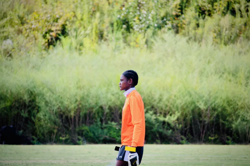 United FC - Furman Region -: A soccer player wearing an orange jersey and goalkeeper gloves stands on a grassy field with tall green foliage in the background.