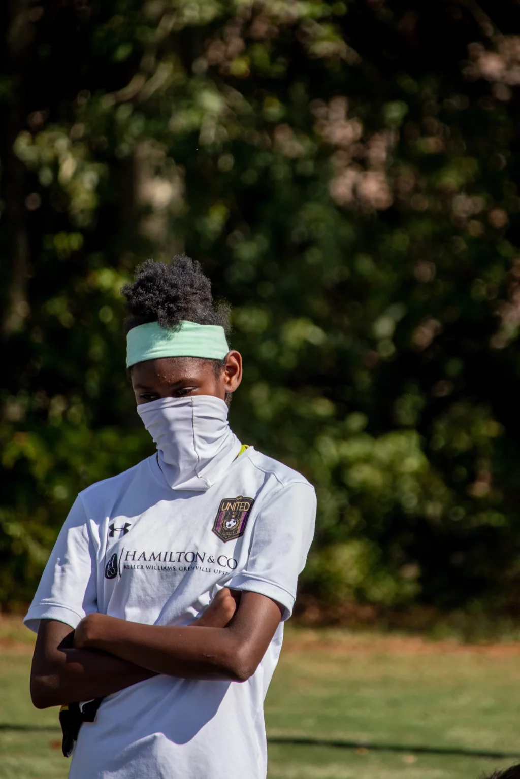 United FC - Furman Region -: A youth soccer player wearing a white jersey, a light blue headband, and a white face covering stands outdoors with arms crossed, with trees blurred in the background.