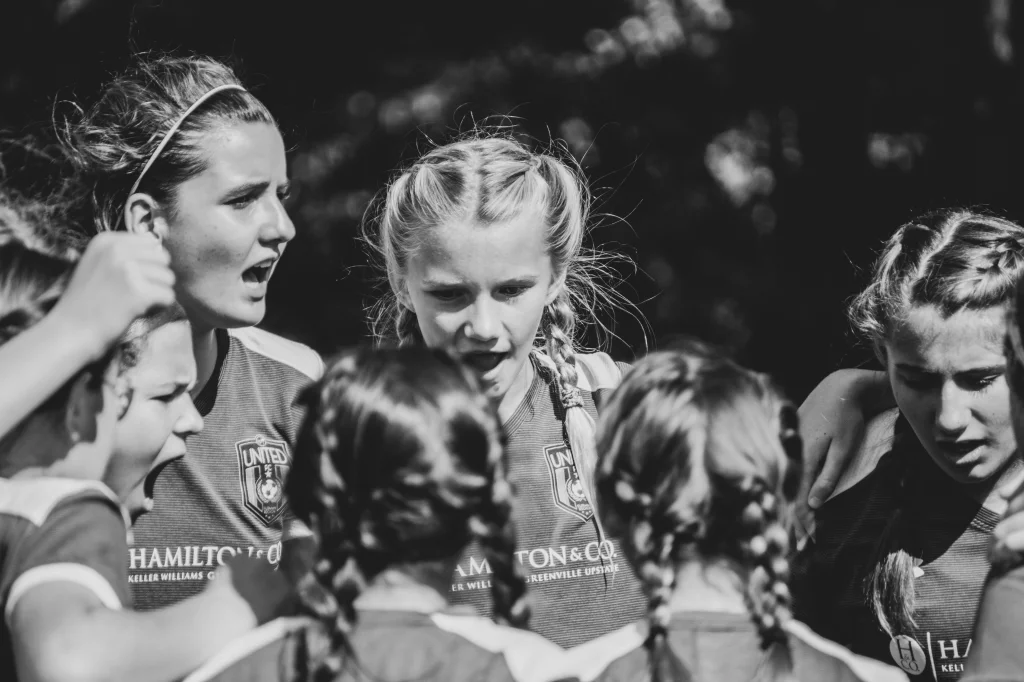 : Black and white photo of a girls&rsquo; soccer team in a huddle, wearing uniforms with braids in their hair, some with intense or focused expressions.