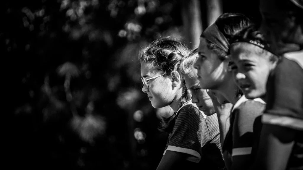: Black and white photo of a group of young girls in striped shirts, sitting in a row and looking intently to the right. The focus is on the girl with glasses, while the others are partially out of focus. The background is blurred.