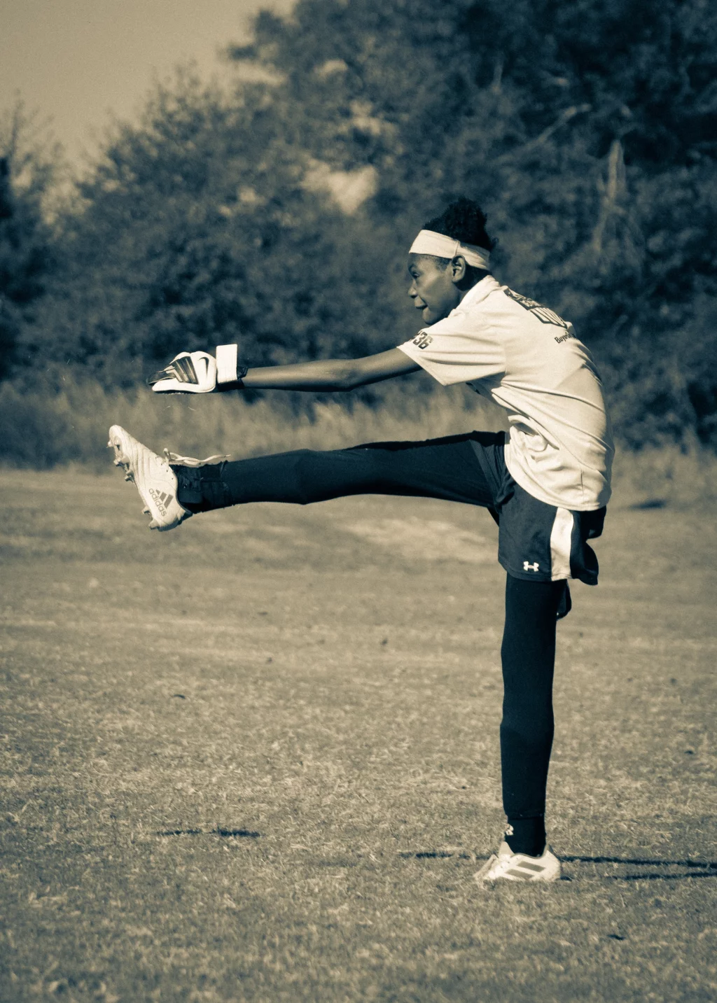 United FC - Furman Region -: A young athlete in sportswear and gloves performs a high kick on a grassy field, with trees in the background. The image is in black and white.