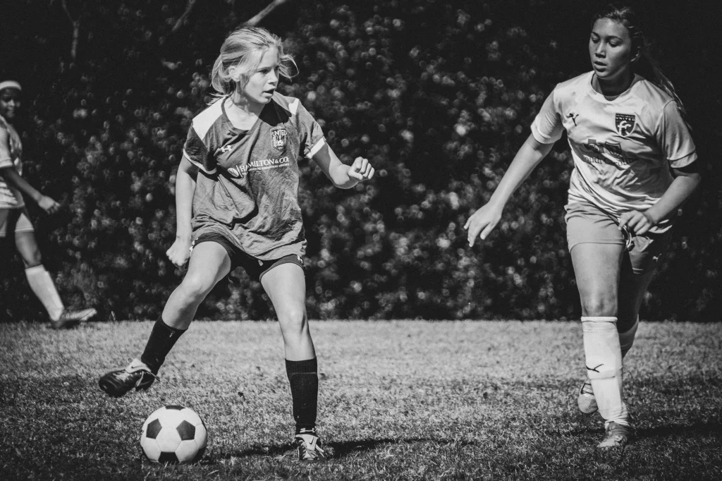: Black and white photo of two girls playing soccer outdoors, one dribbling the ball while the other approaches to defend.